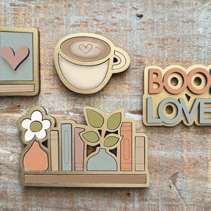 May include: Wooden cutouts of a book with a heart, a coffee cup with a heart, the words "BOOK LOVER", and a bookshelf with books, vases, and a flower. The cutouts are painted in neutral colors with gold accents.