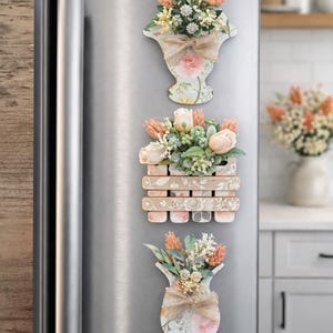 Può includere: Tre magneti decorativi floreali a forma di vaso e cassa sono attaccati a un frigorifero in acciaio inossidabile. I magneti presentano disegni floreali pastello, fiori finti e fiocchi di juta. I fiori hanno accenti pesca, bianchi e verdi.