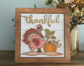 Könnte beinhalten: Ein Schild mit Holzrahmen, weißem Hintergrund und dem Wort "thankful" in goldenen Buchstaben. Das Schild zeigt einen rosa-braunen Truthahn mit einem Kürbis und Blättern.