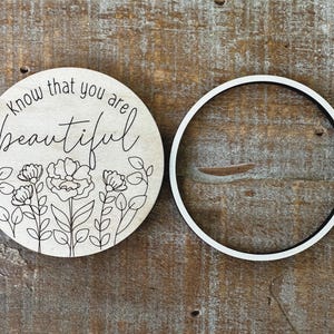 May include: A round wooden sign with the words "know that you are beautiful" in script, along with a floral design. A separate white and black ring is next to the sign.
