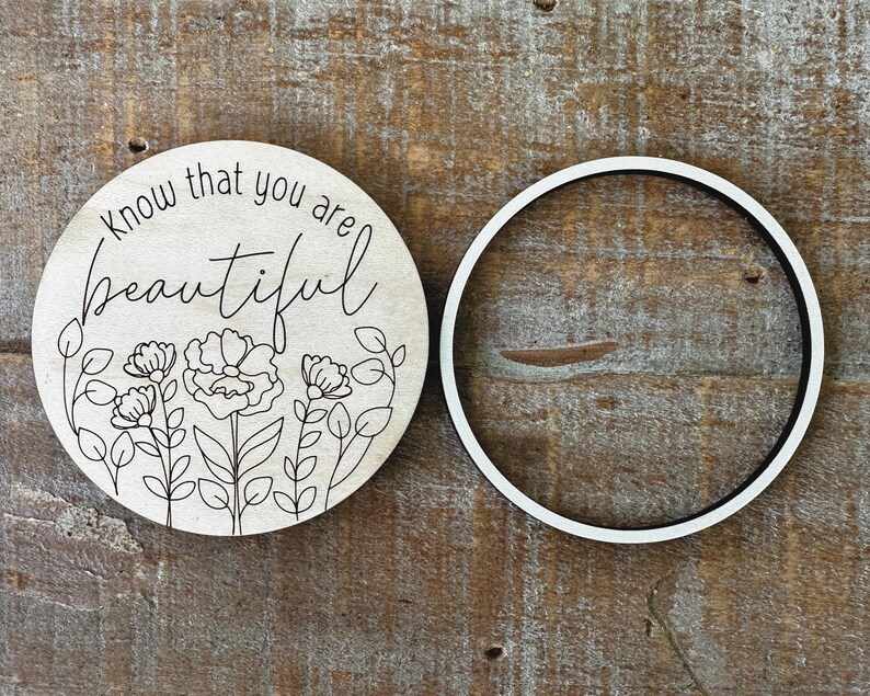 May include: A round wooden sign with the words "know that you are beautiful" in script, along with a floral design. A separate white and black ring is next to the sign.