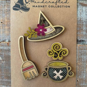 May include: A set of three wooden Halloween magnets featuring a witch's hat with a flower, a cauldron with a skull and crossbones, and a broom. The magnets are handcrafted and have a distressed, vintage look.