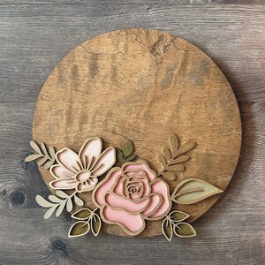 May include: A round wooden sign with a pink and gold floral design. The flowers are layered and have a 3D effect. The sign is perfect for adding a touch of rustic charm to any home.