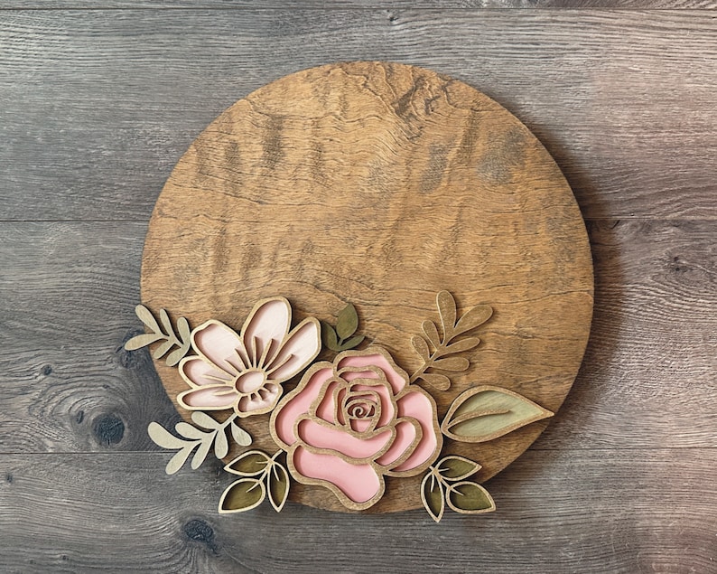 May include: A round wooden sign with a pink and gold floral design. The flowers are layered and have a 3D effect. The sign is perfect for adding a touch of rustic charm to any home.