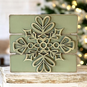 Puede incluir: Un letrero de madera verde con un dise&ntilde;o de copo de nieve de metal dorado. El copo de nieve es intrincado y tiene un dise&ntilde;o delicado.