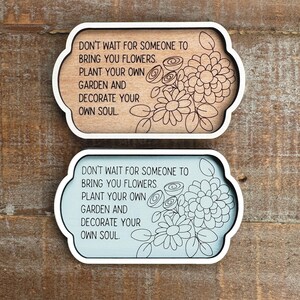 May include: Two decorative wooden signs with a white border and floral design. The signs have the quote, "DON'T WAIT FOR SOMEONE TO BRING YOU FLOWERS. PLANT YOUR OWN GARDEN AND DECORATE YOUR OWN SOUL." One sign is light brown, the other is light blue.