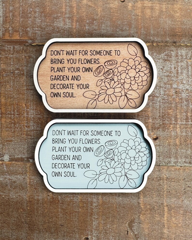 May include: Two decorative wooden signs with a white border and floral design. The signs have the quote, "DON'T WAIT FOR SOMEONE TO BRING YOU FLOWERS. PLANT YOUR OWN GARDEN AND DECORATE YOUR OWN SOUL." One sign is light brown, the other is light blue.