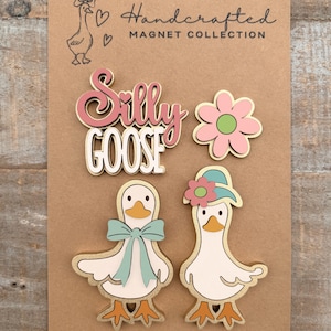 May include: A collection of handcrafted magnets on a brown card. The magnets include a pink and white "Silly Goose" design, a pink flower, and two white geese with gold trim, one with a blue bow and the other with a hat.
