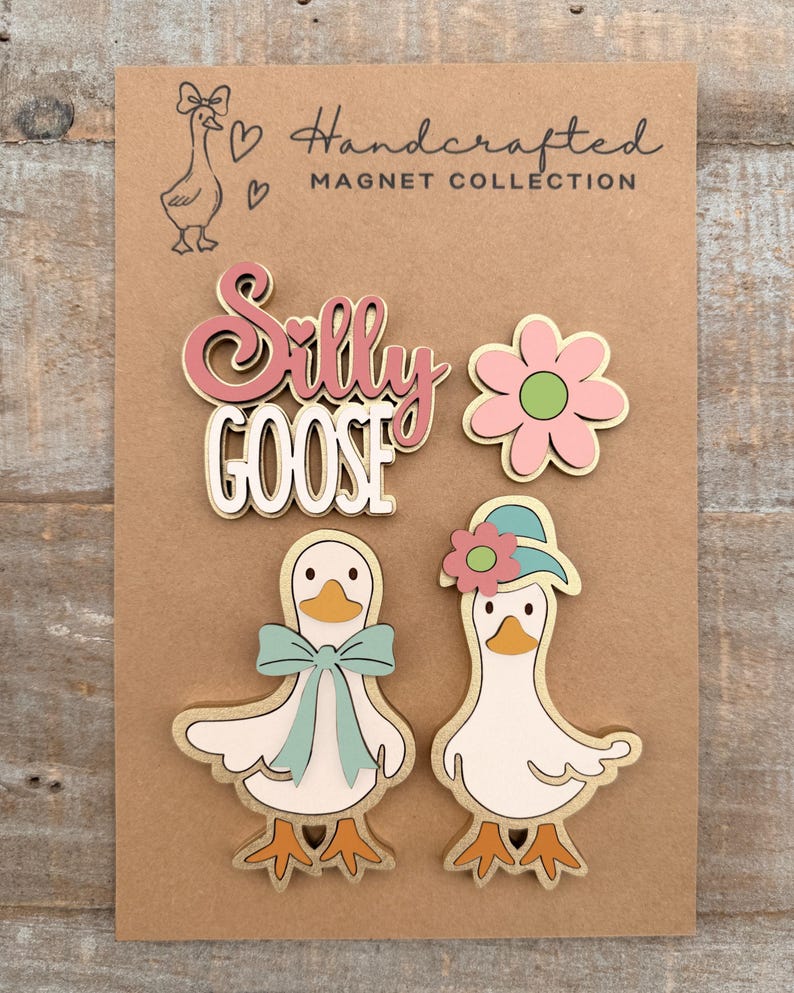 May include: A collection of handcrafted magnets on a brown card. The magnets include a pink and white "Silly Goose" design, a pink flower, and two white geese with gold trim, one with a blue bow and the other with a hat.