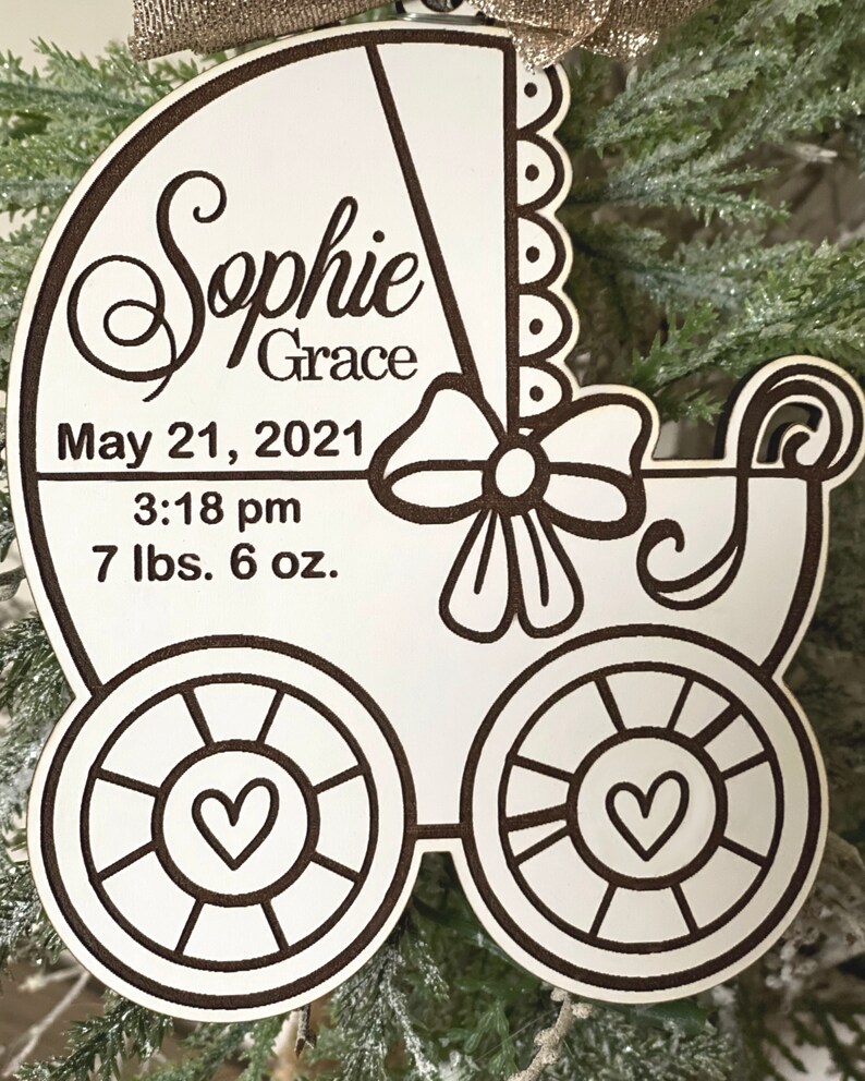 May include: White wooden baby carriage ornament with a black outline. The ornament is personalized with the name "Sophie Grace", the date "May 21, 2021", the time "3:18 pm", and the weight "7 lbs. 6 oz."