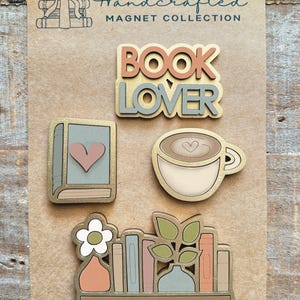 May include: A collection of handcrafted magnets on a brown background. The magnets include the words "BOOK LOVER", a book with a heart, a coffee cup with a heart, and a bookshelf with a flower and books. The magnets have gold trim and pastel colors.