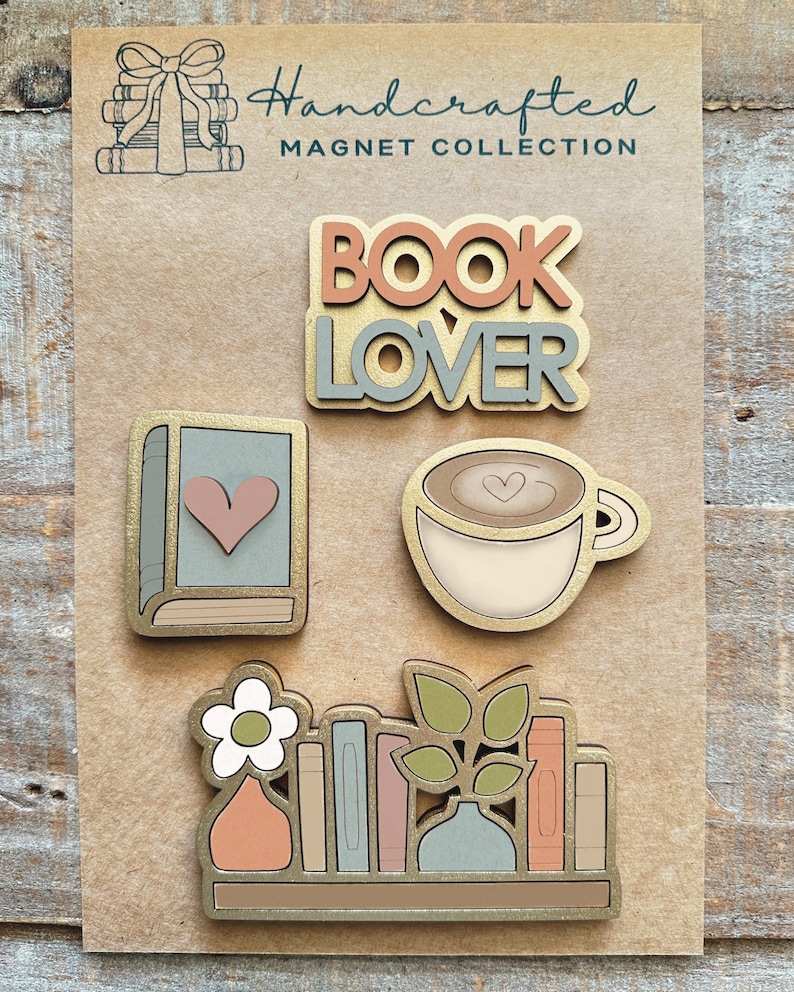 May include: A collection of handcrafted magnets on a brown background. The magnets include the words "BOOK LOVER", a book with a heart, a coffee cup with a heart, and a bookshelf with a flower and books. The magnets have gold trim and pastel colors.