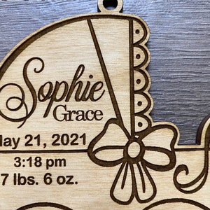May include: A wooden baby carriage ornament with the name "Sophie Grace" engraved on it. The ornament also includes the date "May 21, 2021", the time "3:18 pm", and the baby's weight "7 lbs. 6 oz."