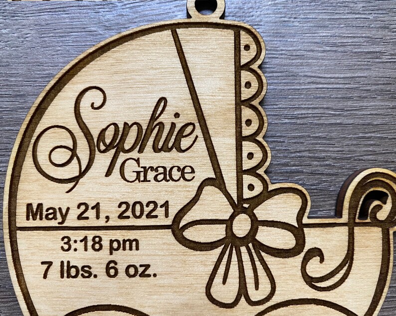 May include: A wooden baby carriage ornament with the name "Sophie Grace" engraved on it. The ornament also includes the date "May 21, 2021", the time "3:18 pm", and the baby's weight "7 lbs. 6 oz."