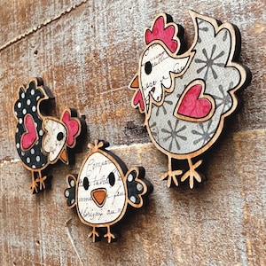 Puede incluir: Tres adornos de pollo de madera con diferentes patrones. Uno es de lunares negros y blancos, otro es gris con copos de nieve blancos y un coraz&oacute;n rojo, y el &uacute;ltimo es blanco con letras negras y grises y un pico rojo.