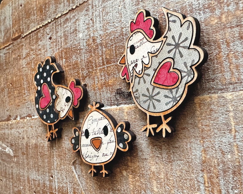 Puede incluir: Tres adornos de pollo de madera con diferentes patrones. Uno es de lunares negros y blancos, otro es gris con copos de nieve blancos y un coraz&oacute;n rojo, y el &uacute;ltimo es blanco con letras negras y grises y un pico rojo.