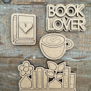 May include: A collection of laser-cut wooden shapes, including a book with a heart, the words "BOOK LOVER", a coffee cup with a heart, and a shelf with books, flowers, and plants. The shapes are light brown and sit on a wooden surface.