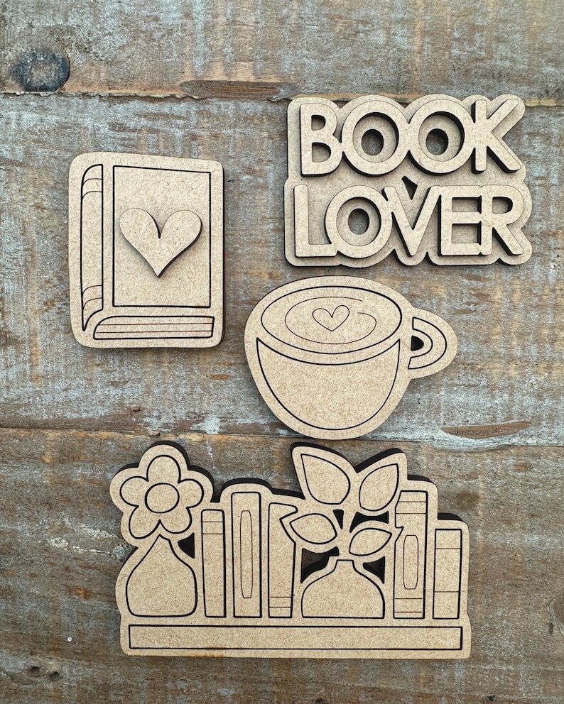 May include: A collection of laser-cut wooden shapes, including a book with a heart, the words "BOOK LOVER", a coffee cup with a heart, and a shelf with books, flowers, and plants. The shapes are light brown and sit on a wooden surface.