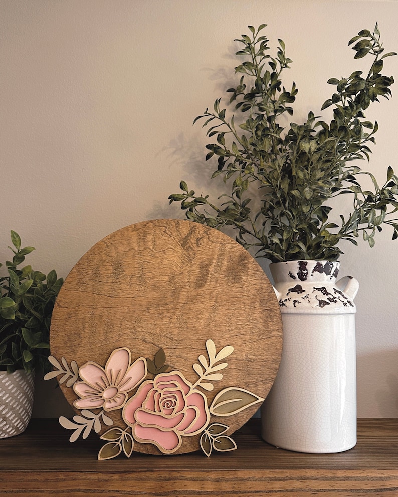 May include: A round wooden sign with a pink and gold floral design. The sign is decorated with a large pink rose and a smaller pink flower with gold leaves.