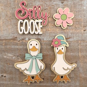 May include: Wooden cutouts of a flower, two geese, and the words "Silly Goose." The geese are white with orange beaks and feet, and one has a blue bow. The other goose wears a hat with a pink flower. The text is in pink and white.