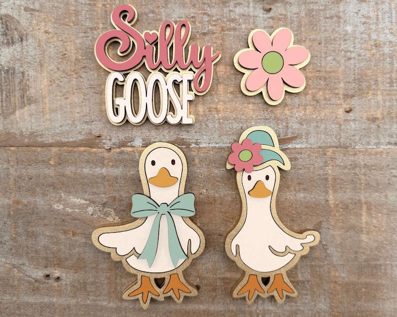 May include: Wooden cutouts of a flower, two geese, and the words "Silly Goose." The geese are white with orange beaks and feet, and one has a blue bow. The other goose wears a hat with a pink flower. The text is in pink and white.