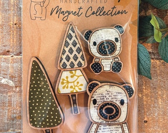 May include: Set of four handcrafted wooden magnets featuring a bear, a tree, and two stylized trees. The magnets are decorated with patterned fabric and printed paper. The text "HANDCRAFTED Magnet Collection" is printed on the packaging.