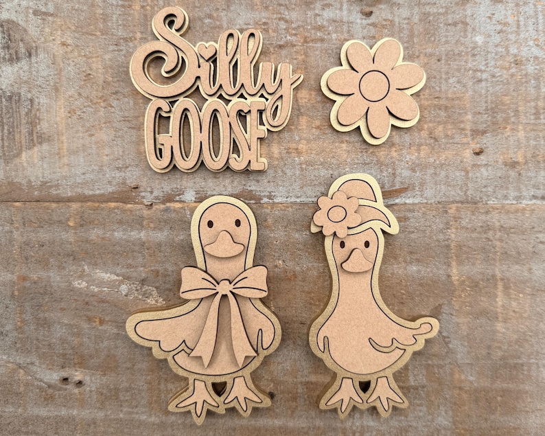 May include: Wooden cutouts of a flower, two geese, and the words "Silly Goose". The geese have bowties and hats. The cutouts are made of light brown wood with a darker brown outline.