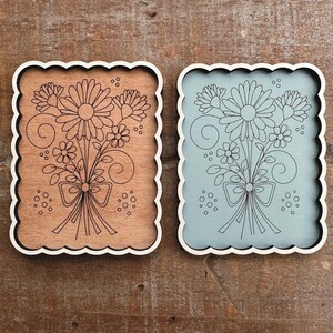 May include: Two decorative wooden plaques with scalloped edges. One is brown with a floral design, the other is light blue. Both feature a black line drawing of a flower bouquet tied with a bow. The plaques are rectangular with rounded corners.