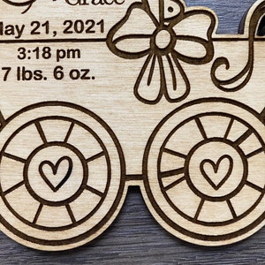 May include: A wooden baby carriage ornament with a bow and hearts. The ornament has the text "May 21, 2021", "3:18 pm", and "7 lbs. 6 oz."