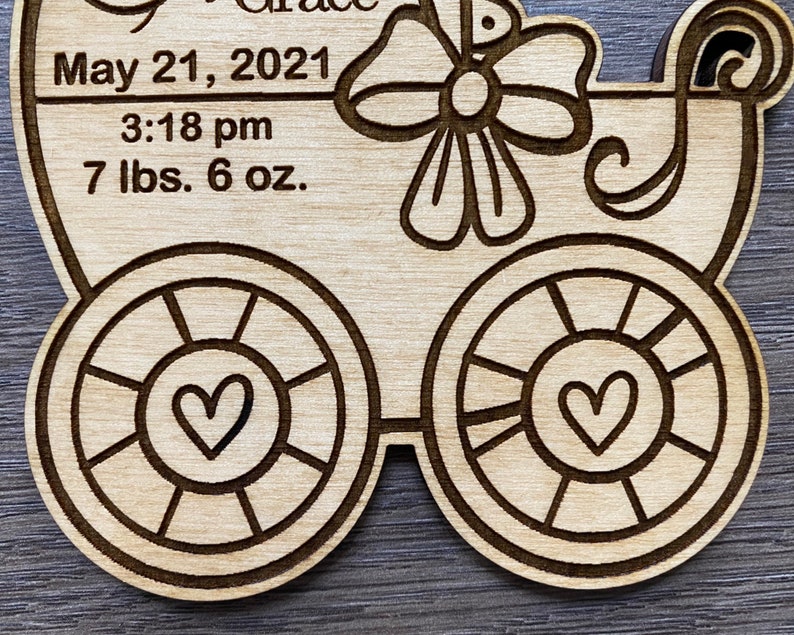 May include: A wooden baby carriage ornament with a bow and hearts. The ornament has the text "May 21, 2021", "3:18 pm", and "7 lbs. 6 oz."