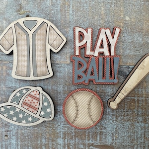 May include: Wooden baseball-themed cutouts, including a jersey, baseball cap, baseball, bat, and the words "PLAY BALL!" The jersey and cap feature star and diamond patterns in red, white, and blue. The baseball has stitched details.
