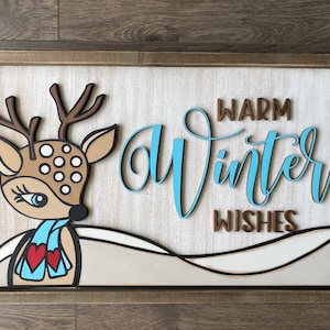 May include: A wooden sign with a brown frame and a white background. The sign features a cutout of a deer with brown antlers, brown spots, and a blue scarf. The text "Warm Winter Wishes" is written in blue and brown letters.