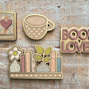 May include: Wooden decorative cutouts featuring a book, coffee cup, the words "BOOK LOVER", and a bookshelf with books and plants. The cutouts have gold edges and pastel colors, with heart and floral patterns.