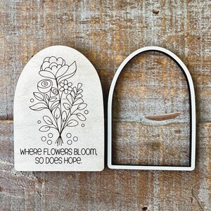 May include: Two wooden arch-shaped cutouts. One features a floral line drawing with the text "WHERE FLOWERS BLOOM, SO DOES HOPE." The other is a white arch-shaped frame with a black interior. The background is a wood grain surface.