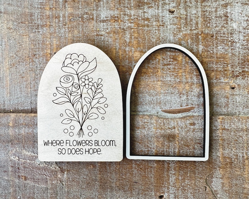May include: Two wooden arch-shaped cutouts. One features a floral line drawing with the text "WHERE FLOWERS BLOOM, SO DOES HOPE." The other is a white arch-shaped frame with a black interior. The background is a wood grain surface.