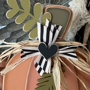 Pode incluir: Uma ab&oacute;bora cor de p&ecirc;ssego com uma fita listrada branca e preta e um cora&ccedil;&atilde;o preto. A ab&oacute;bora &eacute; decorada com folhas verdes e palha.