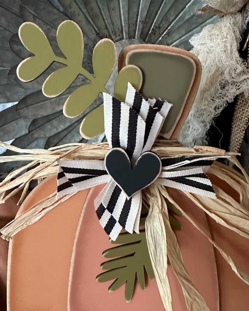 Pode incluir: Uma ab&oacute;bora cor de p&ecirc;ssego com uma fita listrada branca e preta e um cora&ccedil;&atilde;o preto. A ab&oacute;bora &eacute; decorada com folhas verdes e palha.