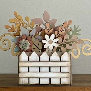 May include: A wooden planter with a picket fence design, filled with a variety of pastel-colored flowers and leaves. The planter is a light brown color with white paint accents.