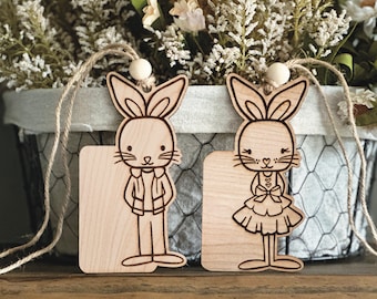 Puede incluir: Dos etiquetas de regalo de madera con forma de conejo con espacios en blanco para escribir. El conejo de la izquierda lleva una chaqueta y pantalones, mientras que el conejo de la derecha lleva un vestido y un lazo. Ambos conejos tienen una cuerda atada en la parte superior.