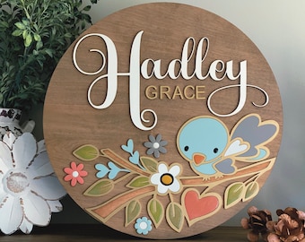 May include: A round wooden sign with the name "Hadley Grace" in white script. The sign is decorated with a blue bird, flowers, and leaves.