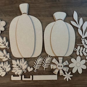 Pode incluir: Um conjunto de recortes de madeira inacabados para artesanato, com duas ab&oacute;boras, folhas, flores e um bot&atilde;o decorativo. As pe&ccedil;as est&atilde;o prontas para serem pintadas, tingidas ou decoradas.