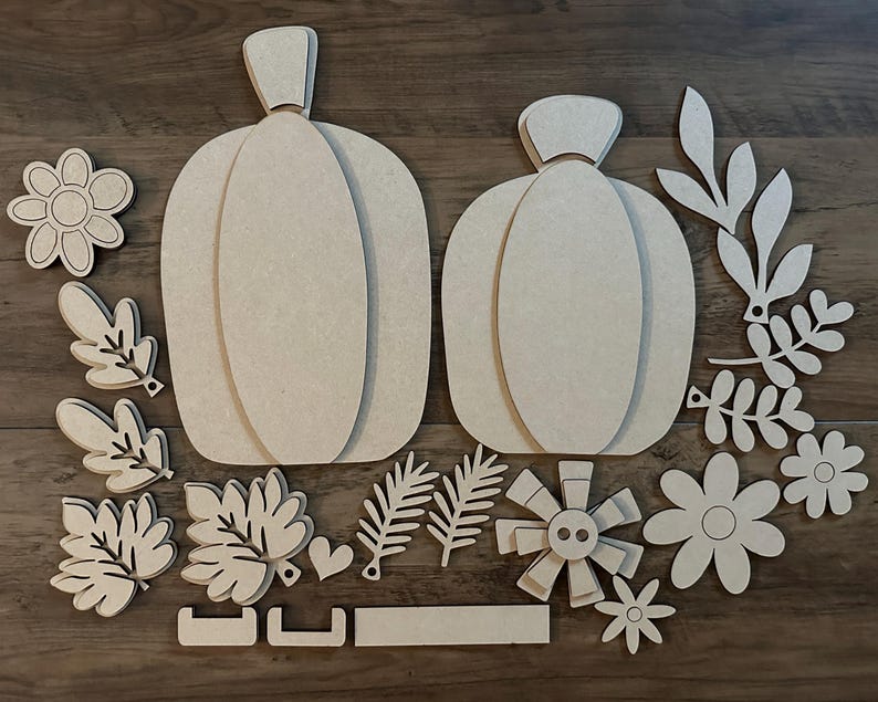 Pode incluir: Um conjunto de recortes de madeira inacabados para artesanato, com duas ab&oacute;boras, folhas, flores e um bot&atilde;o decorativo. As pe&ccedil;as est&atilde;o prontas para serem pintadas, tingidas ou decoradas.