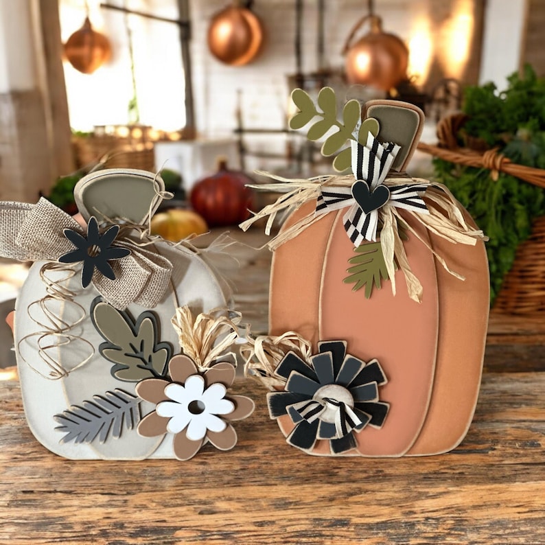 Pode incluir: Duas ab&oacute;boras de madeira decoradas para o outono. A ab&oacute;bora &agrave; esquerda &eacute; branca com folhas e flores marrons, cinzas e verdes. A ab&oacute;bora &agrave; direita &eacute; laranja com uma fita listrada branca e preta, uma flor branca e preta e folhas verdes.