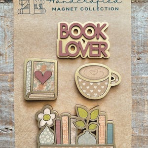 May include: Handcrafted magnet collection featuring a 'Book Lover' design, a book with a heart, a coffee cup with a heart, and a bookshelf with a flower and plant. The magnets have a gold border and are on a brown background.