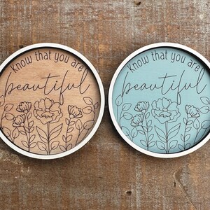May include: Two round wooden signs with white borders. One is light brown with black floral line art and the words "Know that you are beautiful." The other is light blue with the same design.