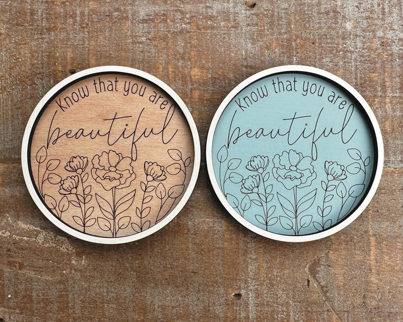 May include: Two round wooden signs with white borders. One is light brown with black floral line art and the words "Know that you are beautiful." The other is light blue with the same design.