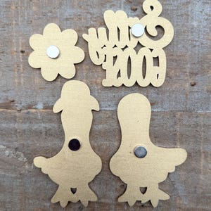 May include: Gold-colored magnets in various shapes: a flower, the word "Mrs", the number "2009", and two bird-like figures. Each magnet has a small, round magnet attached to the back. The items are arranged on a wooden surface.