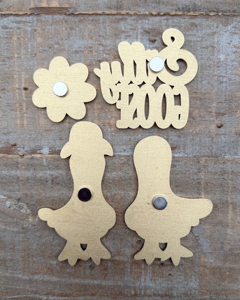 May include: Gold-colored magnets in various shapes: a flower, the word "Mrs", the number "2009", and two bird-like figures. Each magnet has a small, round magnet attached to the back. The items are arranged on a wooden surface.