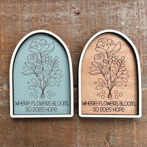 May include: Two wooden plaques with arched tops and white frames. One plaque has a light blue background, the other a natural wood tone. Both feature a floral design and the text "WHERE FLOWERS BLOOM, SO DOES HOPE."
