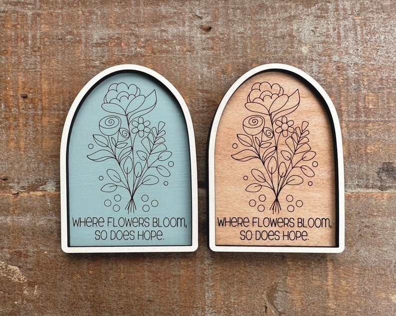 May include: Two wooden plaques with arched tops and white frames. One plaque has a light blue background, the other a natural wood tone. Both feature a floral design and the text "WHERE FLOWERS BLOOM, SO DOES HOPE."